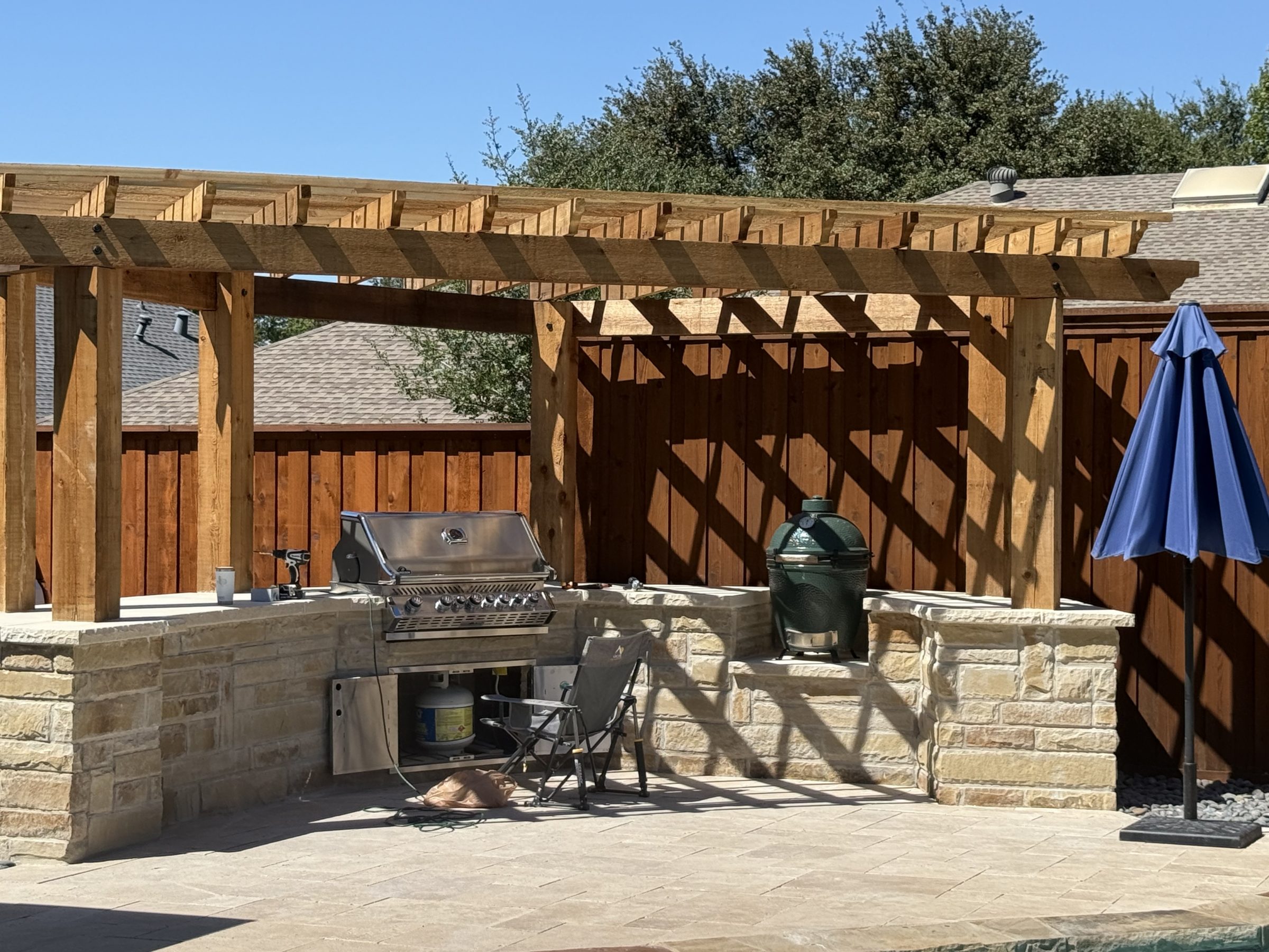Outdoor grilling area with pergola and blue umbrella.